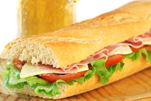 baguette sandwich with lettuce, tomatoes, ham, and cheese with a glass of beer in the background